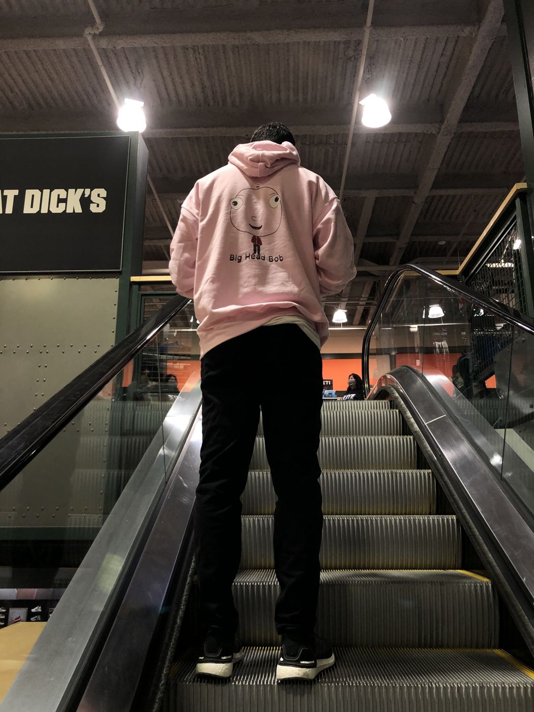 A fan in a pink Big Head Bob hoodie riding a mall escalator